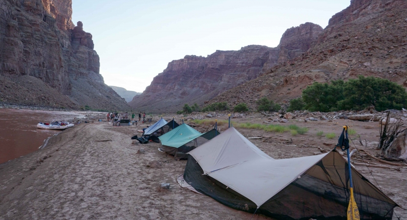 Tarp shelters rest on the bank of a river framed by high canyon walls.
